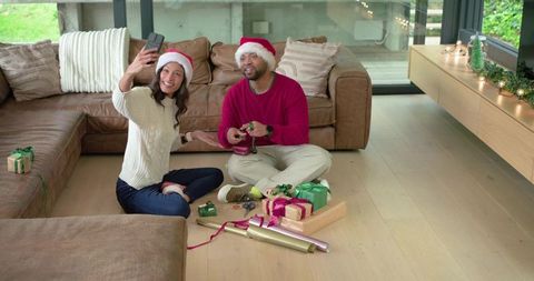 Couple wearing santa hats wrapping gifts and taking selfie in cozy modern living room