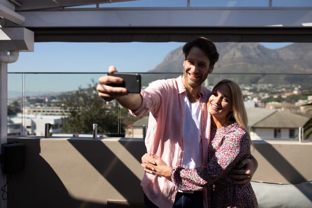 Picture shows a joyful couple embracing and taking a selfie on a sunny rooftop terrace. Ideal for use in advertisements for vacations, hotels, or technology products related to photography and smartphones.