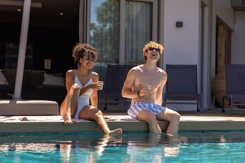 Couple relaxing at poolside holding drinks laughing on modern sunlit patio by pool