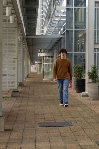 Young man walking under modern glass canopy holding red folder along urban office corridor