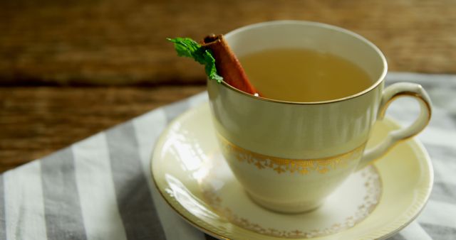 A warm cup of tea with a cinnamon stick and a sprig of mint rests on a saucer atop a striped cloth, with copy space. Its inviting appearance suggests a cozy moment for relaxation or a soothing remedy for cold days.