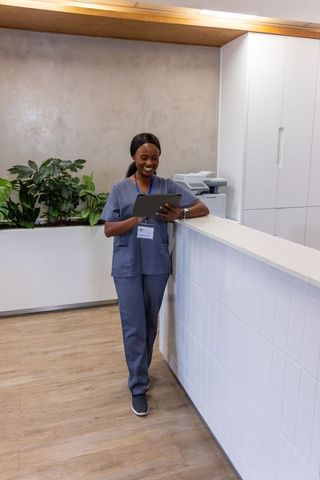 African nurse smiling while using tablet at modern clinic reception desk with plants
