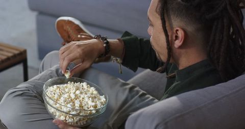 Relaxing man with dreadlocks holding glass bowl of popcorn on gray sofa