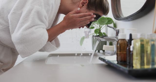 Shows woman washing her face over bathroom sink, engaging in morning skincare routine. Ideal for content on healthcare, self-care routines, beauty tutorials, or skincare product promotions.