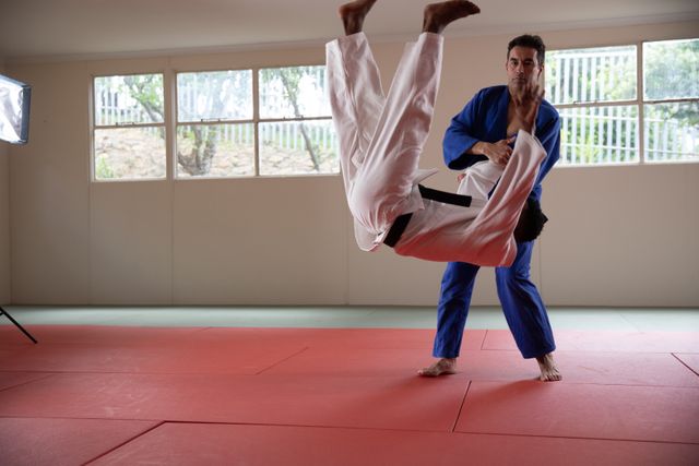Two male judokas, one Caucasian and one African American, are practicing a judo throw on a red mat in a bright studio. The judokas are wearing traditional blue and white judogi. This image can be used for promoting martial arts classes, sports training programs, fitness and self-defense courses, or teamwork and discipline in sports.