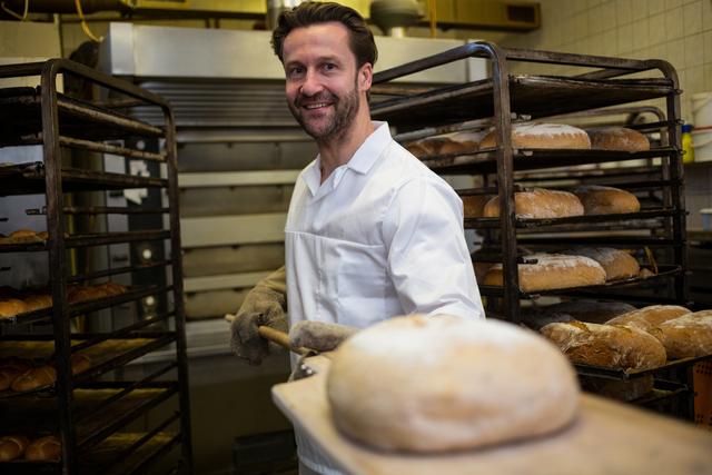 Smiling baker holding freshly baked bread in a bakery. Ideal for use in articles about baking, culinary arts, professional bakers, and bakery businesses. Can be used for promoting bakery products, showcasing baking skills, or illustrating the process of bread making.