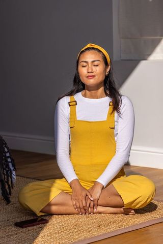 Asian woman meditating in sunlit mustard-yellow overalls practicing mindful breathing