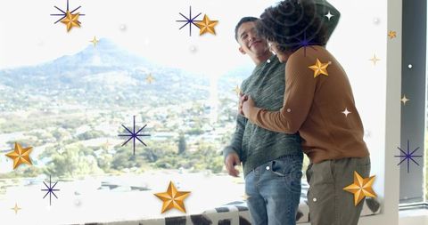 Embracing gay couple leaning by panoramic window overlooking valley, wearing knit sweaters