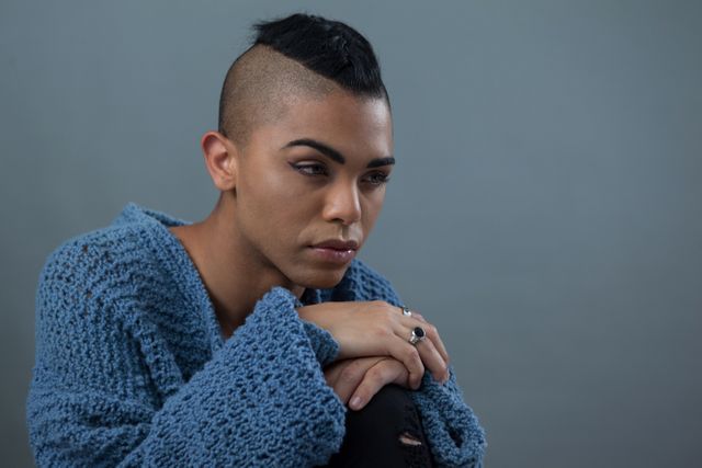 This image depicts a transgender woman with a mohawk hairstyle, wearing a blue sweater, and sitting against a gray background. She appears to be in deep thought, conveying a sense of contemplation and seriousness. This image can be used in articles, blogs, or campaigns focusing on gender identity, LGBTQ issues, individuality, and modern fashion. It is also suitable for use in mental health awareness materials, emphasizing the importance of introspection and self-expression.