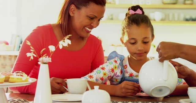 A mother is sitting with her daughter at a breakfast table, both enjoying freshly poured tea. The setting is warm and inviting, indicating a close bond between them. This image can be useful for advertisements, magazines, or websites focusing on family, morning routines, or promoting warm and loving familial relationships. It also works well for content about parenting, breakfast ideas, or daily activities.