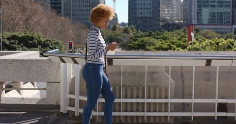 Woman walking on urban pedestrian bridge checking smartphone with city skyline backdrop