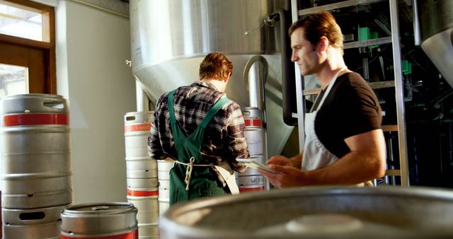 Two men are seen working together in a modern brewery, inspecting large industrial equipment and kegs. They are wearing aprons, indicating their hands-on roles in the brewing process. This image can be used for advertising content in the craft brewing industry, articles on beer production, or visuals for manufacturing and industrial teamwork.