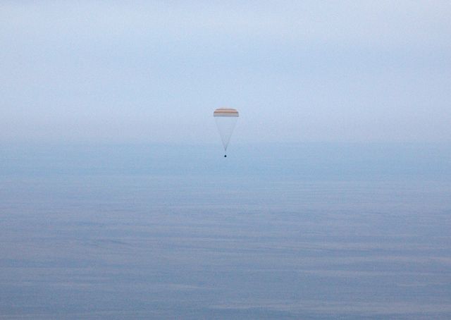 Spacecraft descending with a parachute in open sky. Ideal for use in illustrating space missions, human spaceflight, and international collaborations in space exploration. Could be used in educational materials, news articles, and blogs focused on space programs or historical space missions.