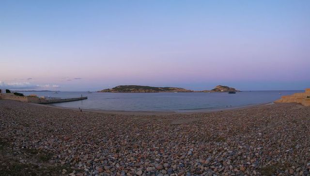 Sunrise pebble beach and calm bay with islands and breakwater under pastel sky