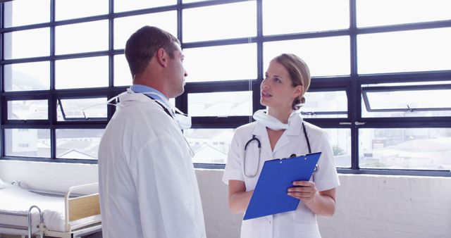 Two doctors standing in a hospital room having a discussion. One doctor holds a blue clipboard, likely discussing patient information or treatment options. Can be used for healthcare, medical teamwork, patient care discussions, or clinical decision-making content.