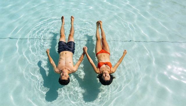 Couple floating and holding hands in sunlit blue pool, overhead, relaxing summer vibe