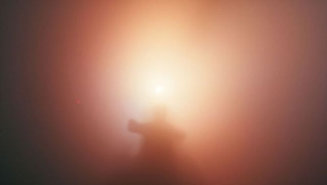 Backlit silhouetted figure reaching through fog toward glowing halo, minimal abstract