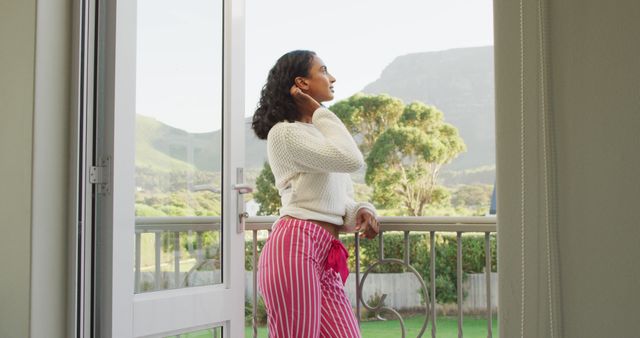 Woman standing on bedroom balcony, enjoying peaceful mountain view. She is wearing a white sweater and red striped pajama pants, reflecting a calm morning atmosphere. Useful for lifestyle blogs, articles about relaxation and mental health, or promotions for travel destinitions and accommodation.
