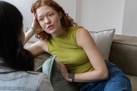 Friends conversing on sofa, relaxed woman reclining with sage cushion and smartwatch