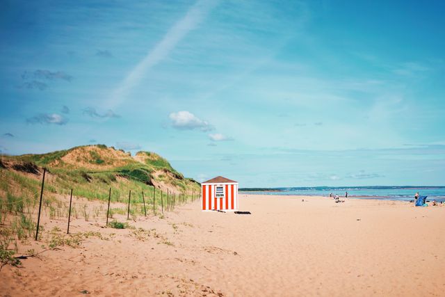 Captures a sunny day on a sandy beach with a distinctive red-and-white striped hut under a clear blue sky. Ideal for illustrating summer vacations, tranquil beachscapes, coastal tourism, travel brochures, or relaxation themes.