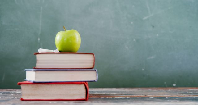 Books stacked on an old school desk with a green apple placed on top, chalk beside, and a backdrop of a blackboard. Ideal for educational materials, school promotions, learning articles, and classroom decor.