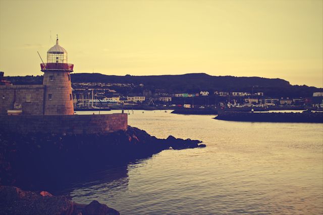 Calm evening at the harbor with a historic lighthouse standing by the waterfront during dusk. The soft colors of the sunset create a serene and peaceful atmosphere. This can be used for travel promotions, coastal scenery backdrops, and thematic content related to maritime navigation.