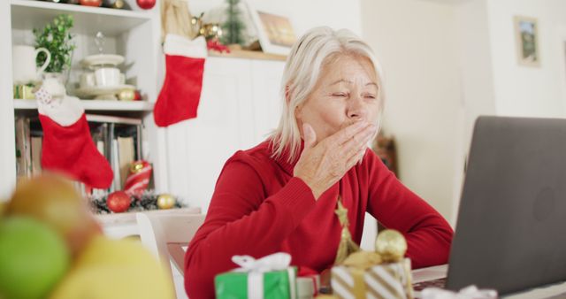 Senior woman, wearing red sweater, blowing kiss towards laptop screen during a virtual Christmas celebration. Festive decorations including stockings, gifts, and ornaments seen in background. Ideal for themes like holiday celebrations, staying connected, family, and senior citizens adapting to technology.