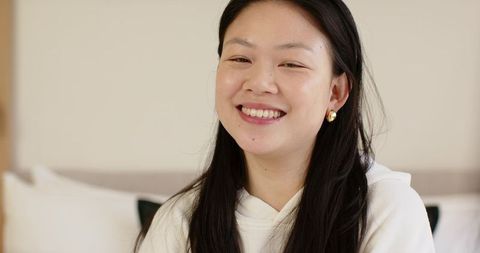 Asian woman smiling while sitting on bed in cozy minimalist bedroom wearing white hoodie