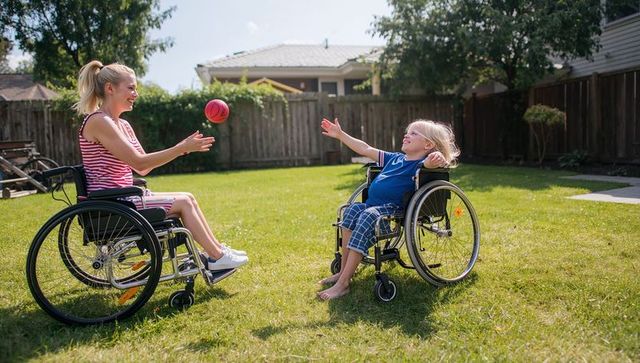 Parent and child in wheelchairs tossing ball on sunny backyard lawn, inclusive playtime