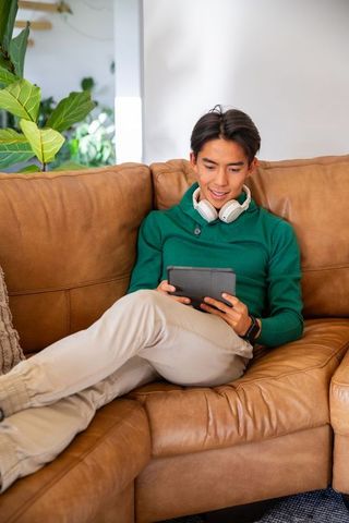 Asian man relaxing on leather sofa, wearing green sweater and headphones, using tablet