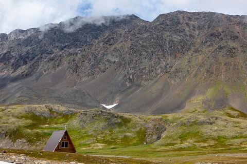 Triangle-shaped cabin nesting beneath rocky mountain in mossy alpine tundra panorama