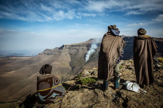 Shepherds dressed in traditional clothing are observing a smoky landscape in the Drakensberg Mountains. They stand and sit on a rugged terrain, reflecting the beauty and harshness of highland life. Ideal for travel and cultural storytelling, highlighting the scenic and cultural richness of the region.