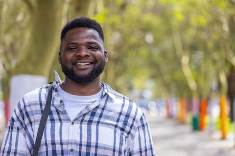 Smiling man standing on sunlit tree-lined path wearing plaid shirt and crossbody bag