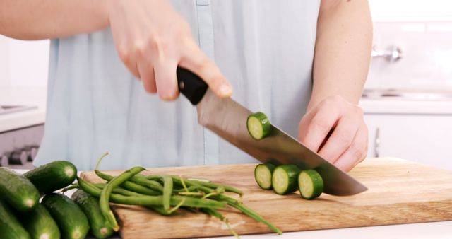 Person chopping fresh cucumbers and green beans on a wooden cutting board with a knife in a modern kitchen. Perfect for use in cooking blogs, healthy eating articles, or meal preparation guides.