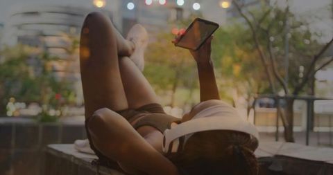 Reclining woman relaxing on rooftop ledge listening to music and scrolling phone at dusk
