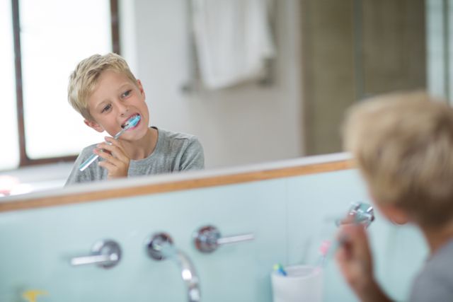 Young boy brushing teeth in front of bathroom mirror, promoting dental hygiene and healthy morning routines. Ideal for use in health and wellness articles, dental care promotions, parenting blogs, and educational materials about personal hygiene.