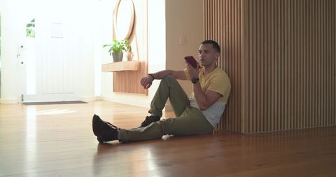 Man relaxing on wooden floor in sunlit modern foyer while using smartphone