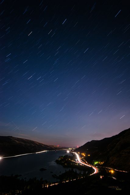 This long exposure photo features a night sky with star trails above a tranquil river. Light trails from vehicles create a luminous path along the winding road in the distance. Illuminated hillsides and faint glowing city lights enhance the serene and picturesque landscape. Ideal for use in travel brochures, nature blogs, photography tutorials, and scenic wallpapers.