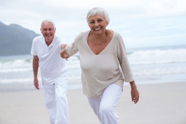 Senior couple smiling and holding hands while enjoying time at the beach. Ideal for use in advertisements promoting retirement plans, senior living communities, travel agencies, and health and wellness products for the elderly.