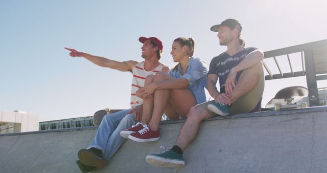 Caucasian woman and two male friends sitting talking and pointing something in distance on sunny day. hanging out at skatepark in summer.