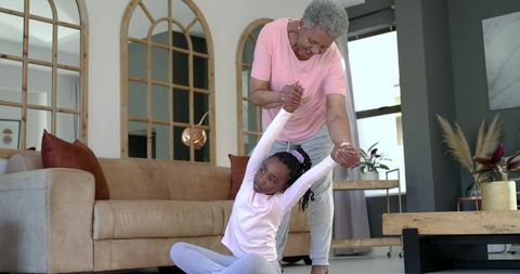Black grandmother guiding granddaughter in gentle seated stretch at home, family bonding