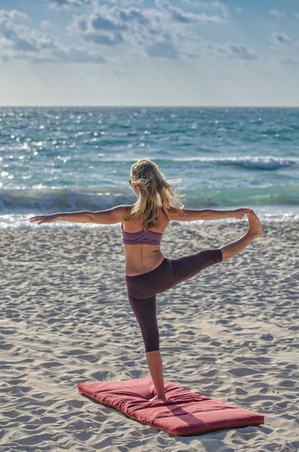 Blonde woman practicing yoga by oceanfront, balancing on one leg on sandy beach during sunset. Ideal for promoting wellness, fitness routines, serene landscapes, and outdoor exercises. Perfect for use in articles, advertisements, websites related to yoga, health and relaxation.
