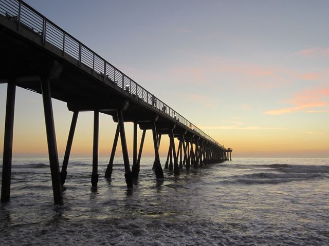 This striking image captures a tranquil sunset view of an ocean pier extending into the calm waters. The soft hues of the sky contrast with the dark silhouette of the pier, creating a serene and peaceful scene perfect for themes around travel, relaxation, and natural beauty. This visual can be used for websites, blogs, travel magazines, or any project looking to evoke a sense of calm and serenity, ideal for promoting coastal destinations and outdoor recreation.
