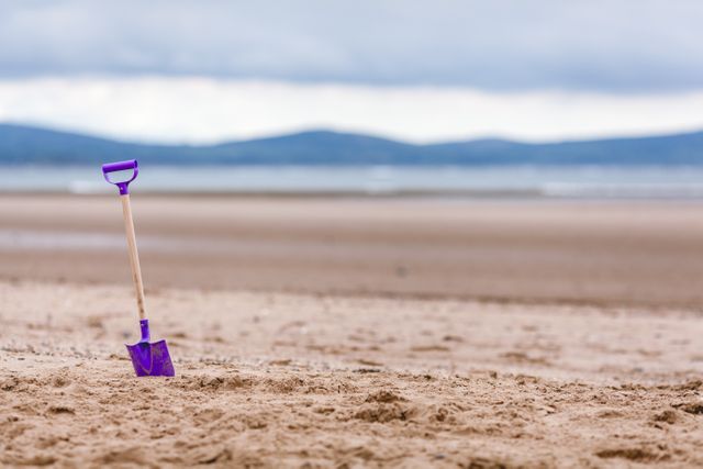 Shovel standing upright in sandy beach near ocean on cloudy day. Suitable for illustrating beach trips, childhood playtime, and vacations. Good for use in travel blogs, promotional content for beach destinations, and advertisements related to outdoor activities.