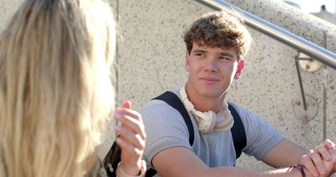 Smiling male student wearing headphones chatting with friend on sunlit campus steps