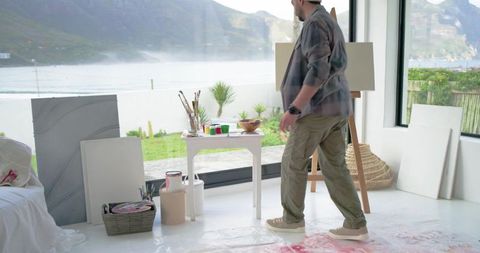 Indian artist walking toward easel in bright coastal studio with seaview and canvases