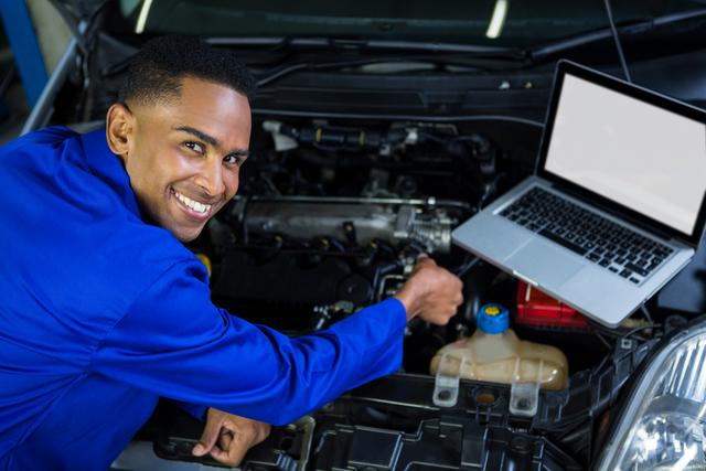 Mechanic in blue uniform smiling while using a laptop to inspect a car engine in a repair garage. Ideal for use in articles or advertisements related to automotive repair, technology in car maintenance, professional services, and workshops.