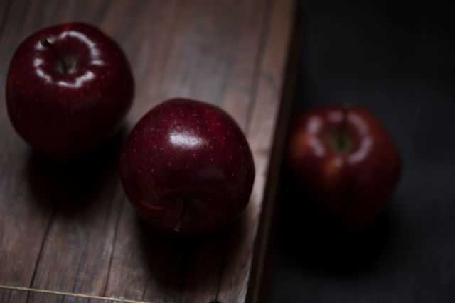 Depicting three fresh organic red apples on a dark wooden surface, this image captures the simplicity and natural beauty of fresh produce. The low light and natural shadow create a rustic and moody aesthetic. Suitable for use in organic food promotions, healthy eating campaigns, or autumn-themed content. Perfect for blog posts, recipe illustrations, and eco-friendly advertisements.