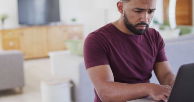 Young man working on laptop in a modern living room environment. Ideal for illustrating concepts of remote work, freelancing, home office setup, and modern technology use in everyday life settings.