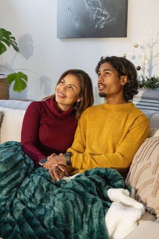 Diverse couple cuddling on couch holding hands under plush blanket, cozy winter evenings